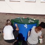 Studnia w kolorze niebieskim i zielonym wiekiem. Przy niej siedzą dwie dziewczyny, które malują na niej wzory,