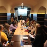 Szkolenie. Podłużna sala, na środku stół przy którym siedzi 25 osób. W tle ekran, a przed nim stoi prowadząca szkolenie.