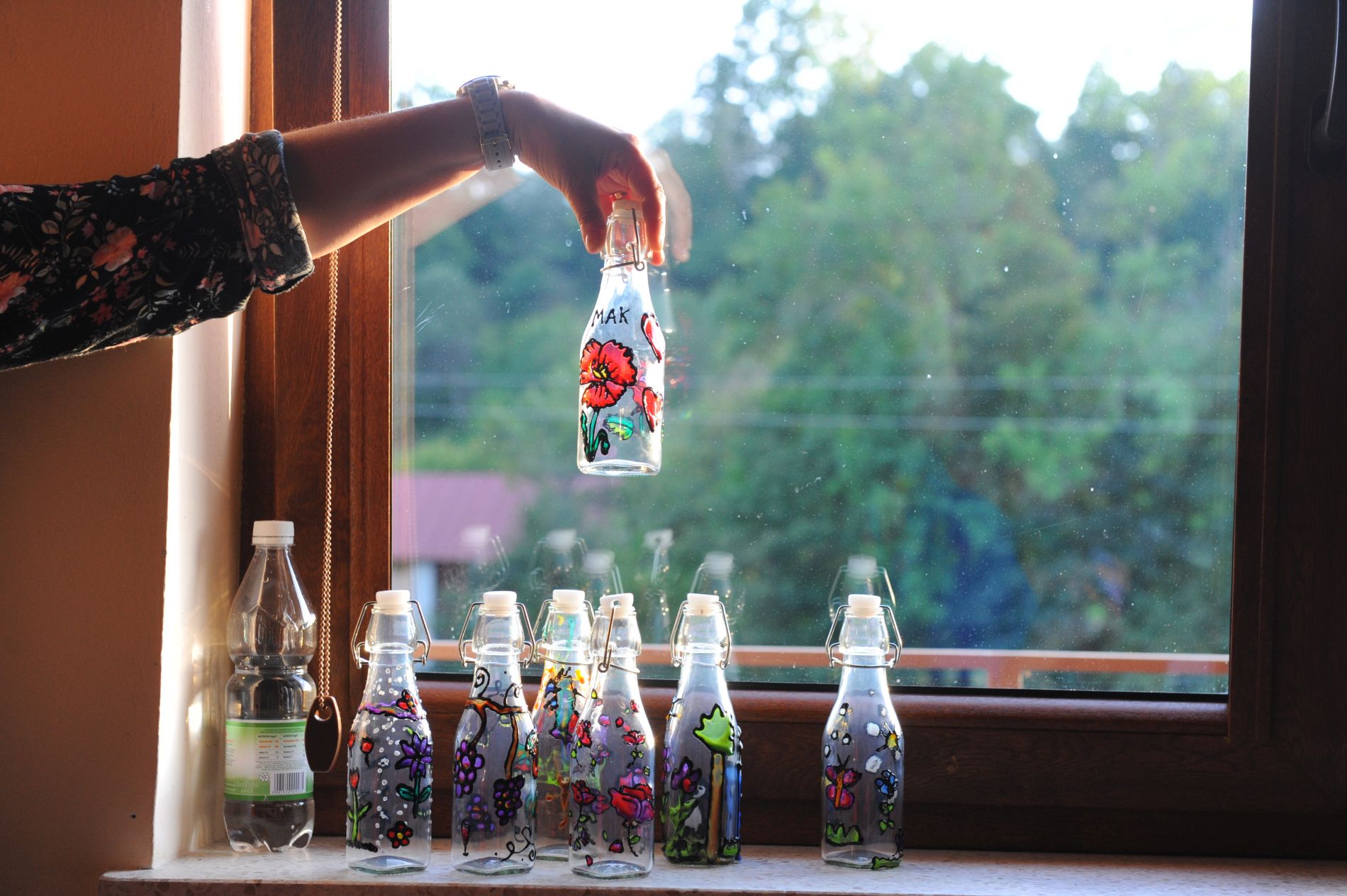 W tle okno. Na parapecie stoi sześć przeźroczystych szklanych butelek z namalowanymi kwiatami, obok plastikowa butelka z wodą mineralną. Na tym wszystkim jedna szklana butelka z namalowanymi kwiatami trzymana w powietrzu w ręce.