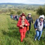 Grupa 11 osób biorących udział w rajdzie, idących pod górę. Na przedzie trzy seniorki. W tle łąki i pagórki.