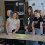 Trójka dziewczyn stojących za stołem. W tle dwa roll-upy: Radia Rzeszów i Bardzo Młodej Kultury. Na stole trzy trójkąty z papieru z żółtą obwolutą.