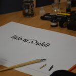 Kartka papieru leżąca na biurku. Na kartce napis "lato w Dukli" pismem kaligraficznym.