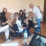 Na pierwszym planie laptop na stoliku. Na drugim planie młodzież siedzi na krzesłach z kartonowymi pudełkami, nad nimi pochyla się instruktor fotografii