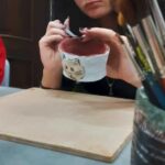 Na pierwszym planie fragment kubek z pędzlami. Na drugim dziewczyna w okularach malująca pędzlem ceramiczny kubek