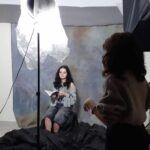 Atelier fotograficzne. Z lewej strony stoi fotograf, z prawej dziewczyna - uczestniczka warsztatów. Na drugim planie siedzi dziewczyna z książką w ręce, za nią czarne płótno. Pośrodku stoi duża lampa fotograficzna.
