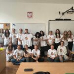 Grupa dziewczyn stojąca w klasie szkolnej, na tle tablicy, w ręce trzymają białe koszulki BMK
