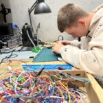 Stół, na pierwszym planie plątanina kabli, nieco dalej nad stołem pochylony chłopak z miernikiem napięcia