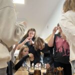 Stoliki ze szklanymi butelkami, nad nim pochylają się dziewczyny, niektóre wąchają zawartość butelek
