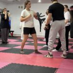 Na pierwszym tle dziewczyna z instruktorem trenują samoobronę. W tle grupa pozostałych ćwiczących dziewczyn