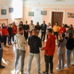Grupa młodzieży stojąca w kółku, w sali szkolnej, pośród nich prowadząca warsztaty.