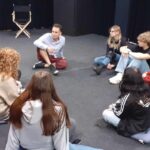 Grupa młodzieży siedząca na dywanowej podłodze, wśród nich instruktor teatralny.